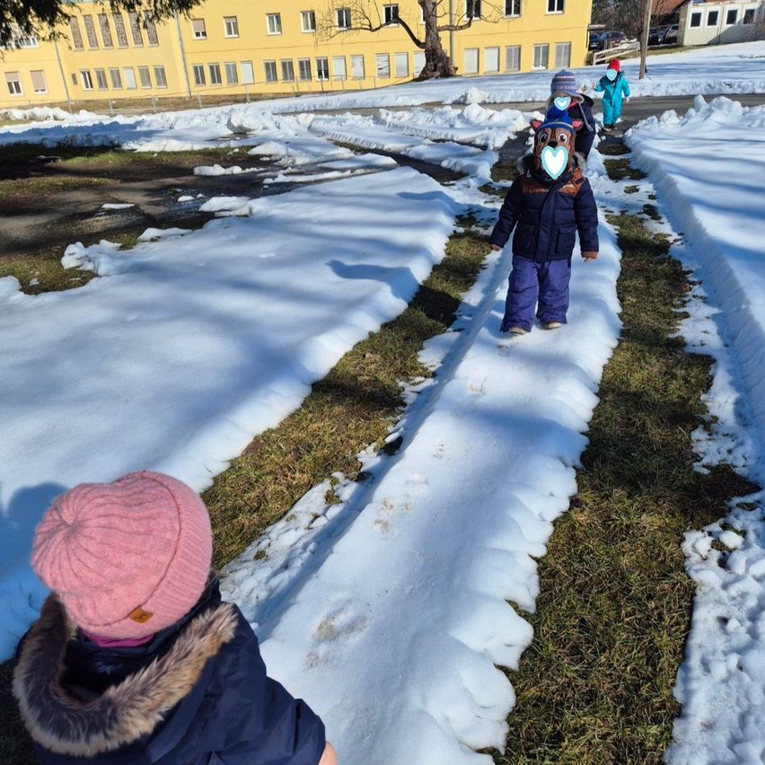Several children walk on snow-covered ground, wearing winter clothes and hats. One child wears a mask. Behind them, there is a building with yellow walls.