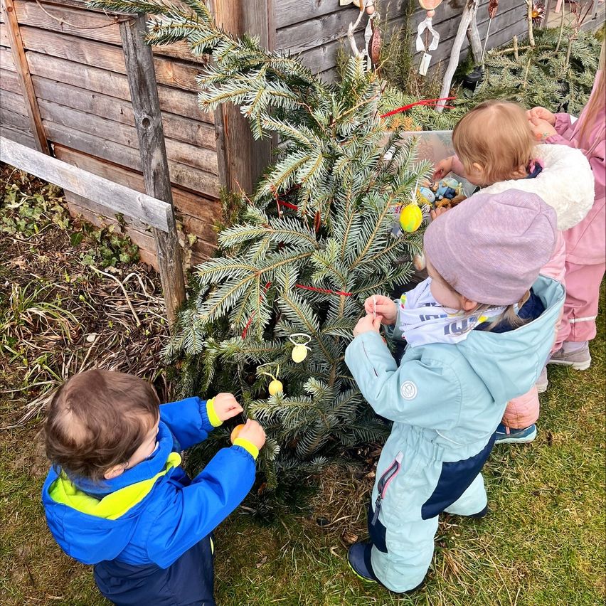 Several children are decorating a tree with ornaments. They are wearing winter clothes and some have hats. A wooden structure is behind them.