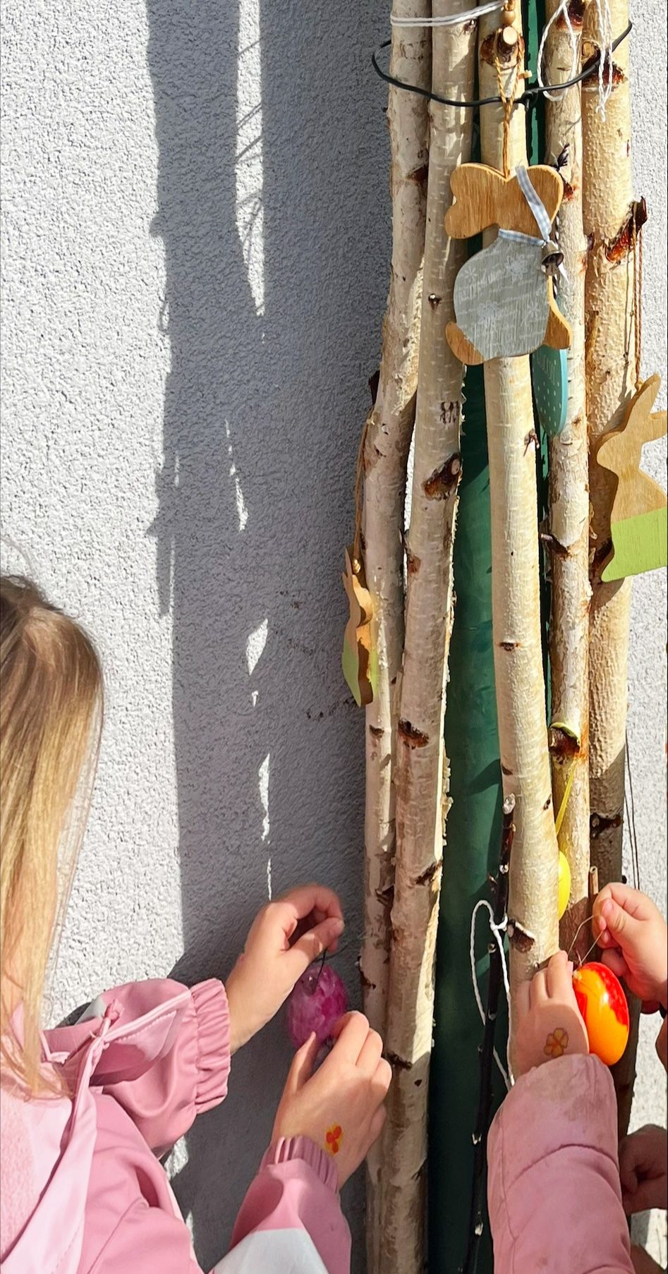 A girl decorates a tree branch arrangement on a wall with eggs and wooden animals. She is seen from behind, with her shadow cast on the wall.