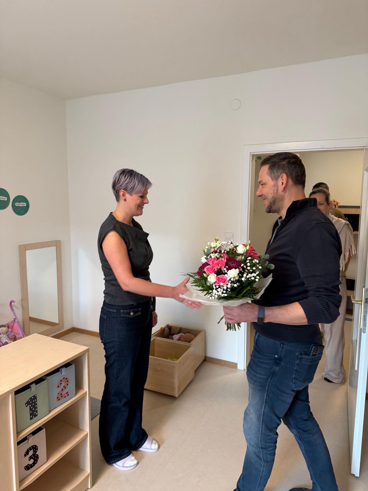 Eine Frau mit lila Haaren überreicht einem lächelnden Mann einen Blumenstrauß in einem Raum mit weißen Wänden und einer Holzkiste mit Spielzeug.