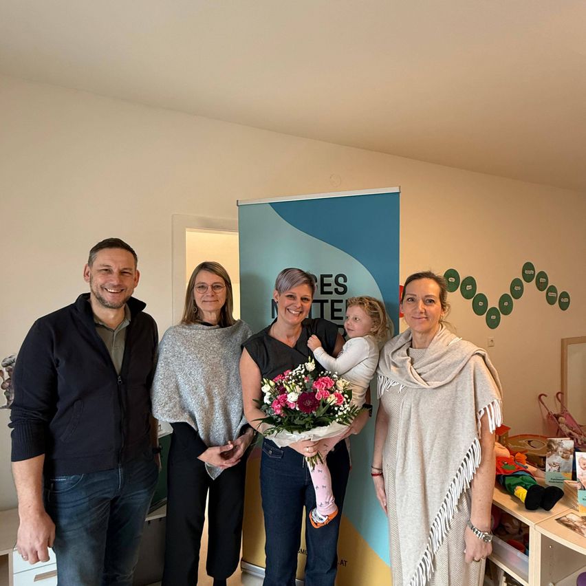 Eine Gruppe von vier Personen steht in einem Raum mit einem Banner hinter ihnen. Eine Frau hält einen Blumenstrauß und ein Baby. Der Mann auf der linken Seite lächelt, während die Frau auf der rechten Seite in die Kamera schaut.