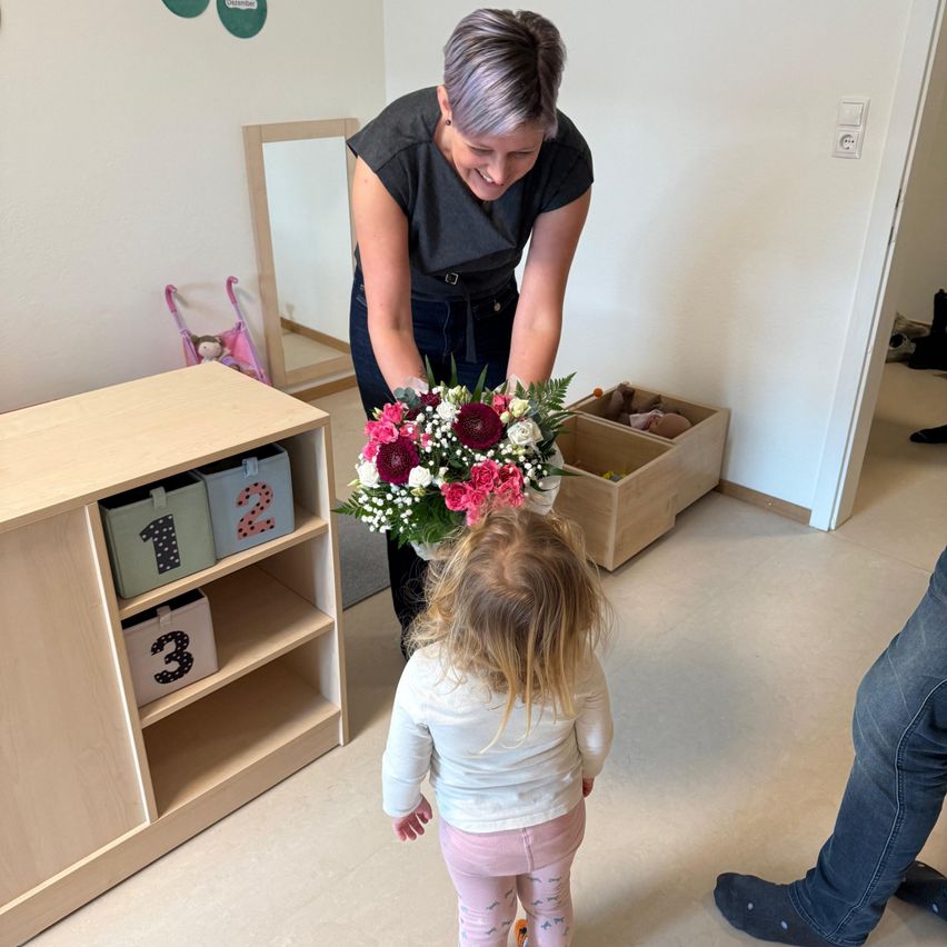 Eine Frau überreicht einem kleinen Mädchen einen bunten Blumenstrauß, das einem Schrank mit nummerierten Boxen gegenübersteht. Ein Mann steht hinter dem Mädchen.