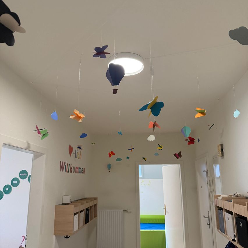 Ein Flur mit weißen Wänden und verschiedenen farbigen Papierdekorationen an der Decke. An der Wand befindet sich ein Schild mit der Aufschrift 'Willkommen' und eine Lampe darüber. Ein Holzregal mit Aufbewahrungsboxen und einem weißen Objekt.