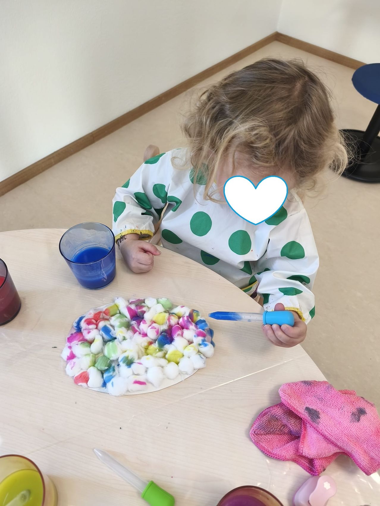 Ein kleines Mädchen sitzt an einem Tisch, hält eine Pipette und ist dabei, blaue Flüssigkeit über eine bunte Baumwollkreation zu gießen. Sie trägt ein grün gepunktetes Lätzchen.