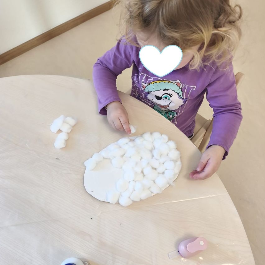 Ein kleines Mädchen sitzt mit einem Bastelprojekt am Tisch. Sie trägt ein lila Hemd und hat Wattebällchen in den Händen. Der Tisch hat ein weißes Papier und einen Herzsticker.