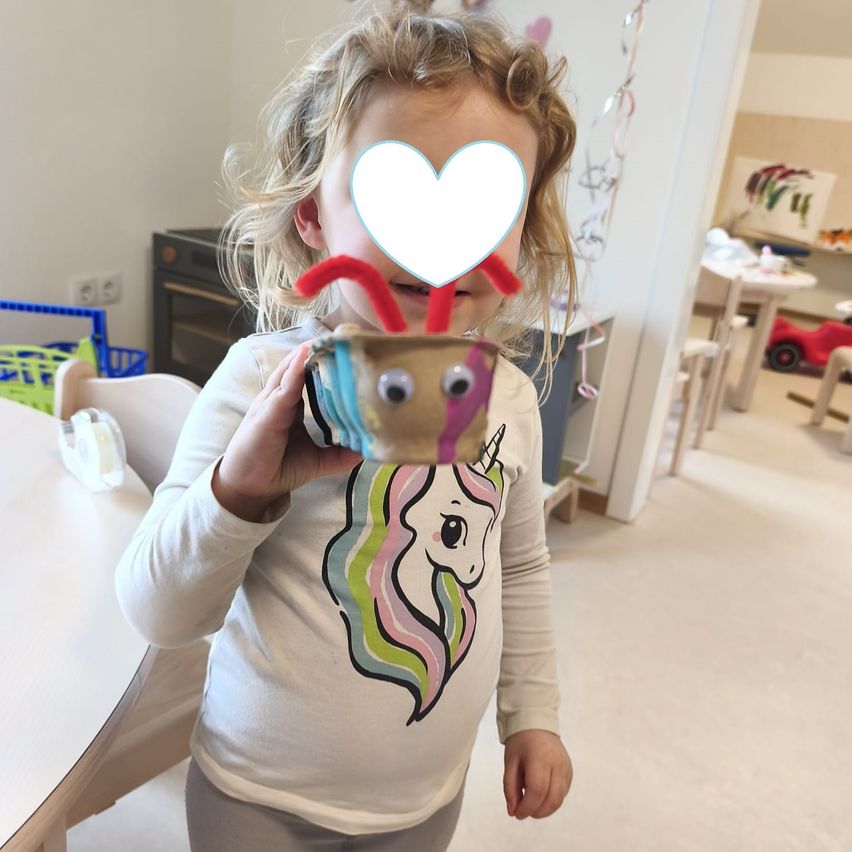 A young girl with blonde hair is holding a crafted object resembling a bee. She is wearing a unicorn-themed long-sleeved shirt. In the background, there is a room with a white wall and a red toy car.