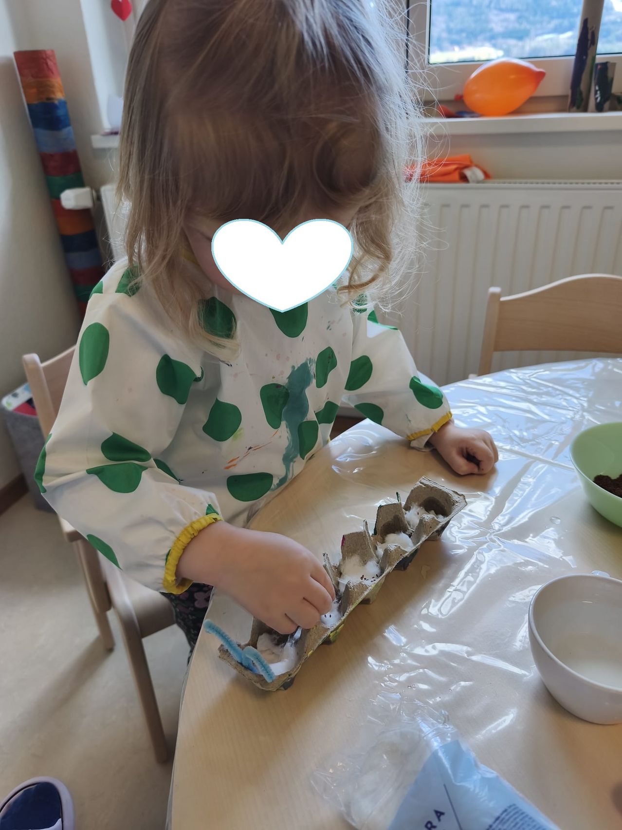 A young girl is sitting at a table, working with an egg carton. She is wearing a green-dotted apron.