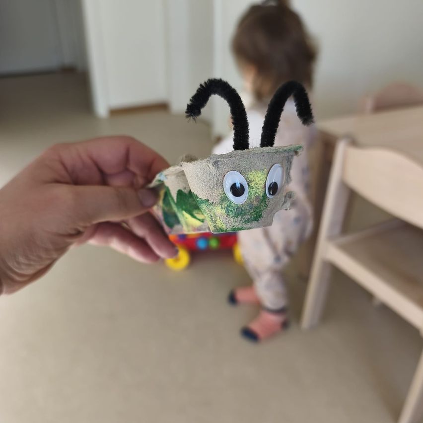 A person holds a homemade caterpillar craft in a living room. A child stands nearby, and a wooden chair is visible.