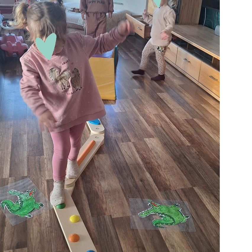Ein kleines Mädchen läuft auf einem Balancebalken mit Socken an den Füßen, mit einem Krokodilsticker auf dem Boden. Ein zweites Mädchen läuft hinter ihr.