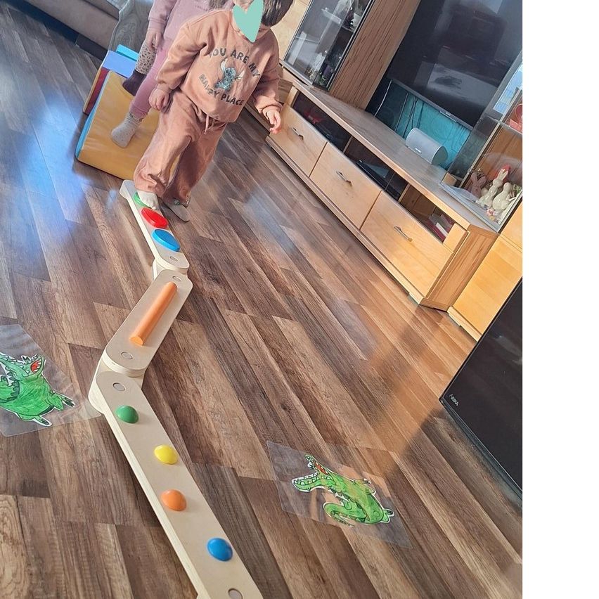 Zwei Kinder spielen mit einem Holzlabyrinth auf dem Boden, eines tritt auf die Knöpfe. Das Labyrinth hat bunte Kreise und Aufkleber eines Krokodils sind auf dem Boden platziert.