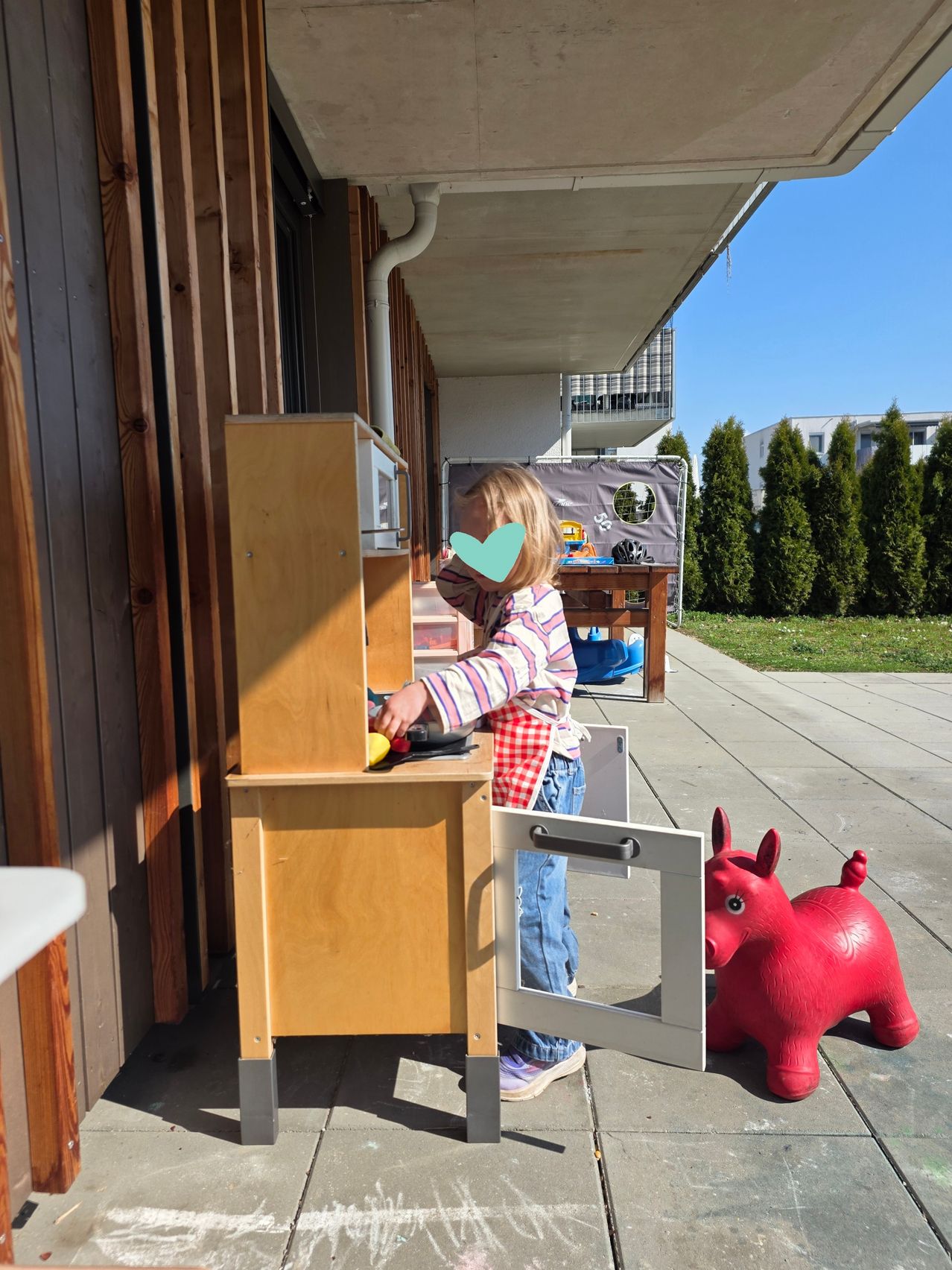 Ein kleines Mädchen spielt in einer Spielküche auf einer Veranda mit einem roten Spielzeugtier in der Nähe.