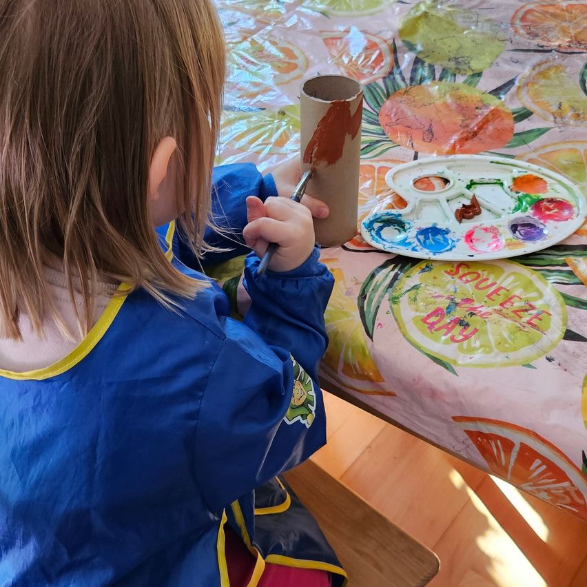 Ein Kind, das einen blauen Malkittel trägt, malt mit einem Pinsel eine braune Tube. Die Malpalette steht auf einer Tischdecke mit Fruchtdesign.