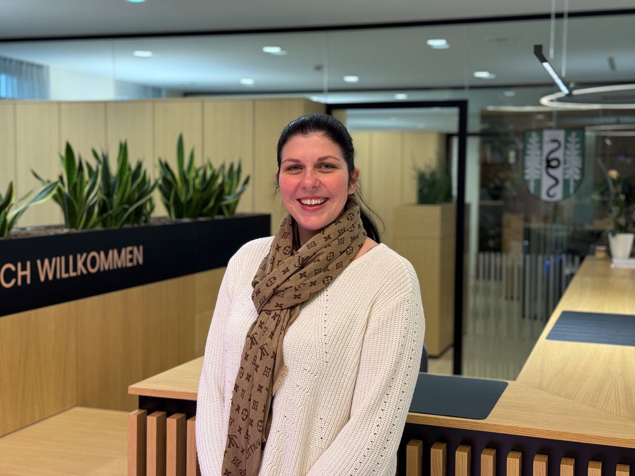 Eine lächelnde Frau in einem weißen Pullover steht in einem Büro und trägt einen Schal. Hinter ihr sind Holztrennwände und grüne Pflanzen zu sehen. Das Büro verfügt über moderne Beleuchtung und eine saubere, professionelle Atmosphäre.