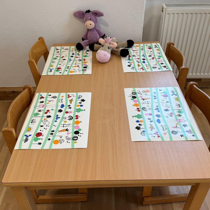 Ein hölzener Tisch für vier Personen mit farbenfrohen Platzsets. Zwei Kuscheltiere liegen auf dem Tisch, und ein weißer Heizkörper ist an der Wand.