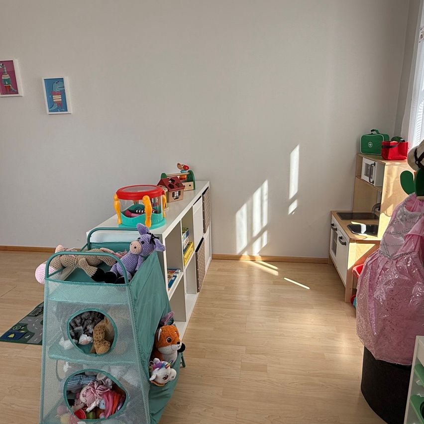 Ein Zimmer mit weißen Wänden, Holzböden und einem weißen Regal voller Spielzeug. Zwei Bilder hängen an der Wand. Spielzeug und ein Spielzeugorganizer liegen auf dem Boden.