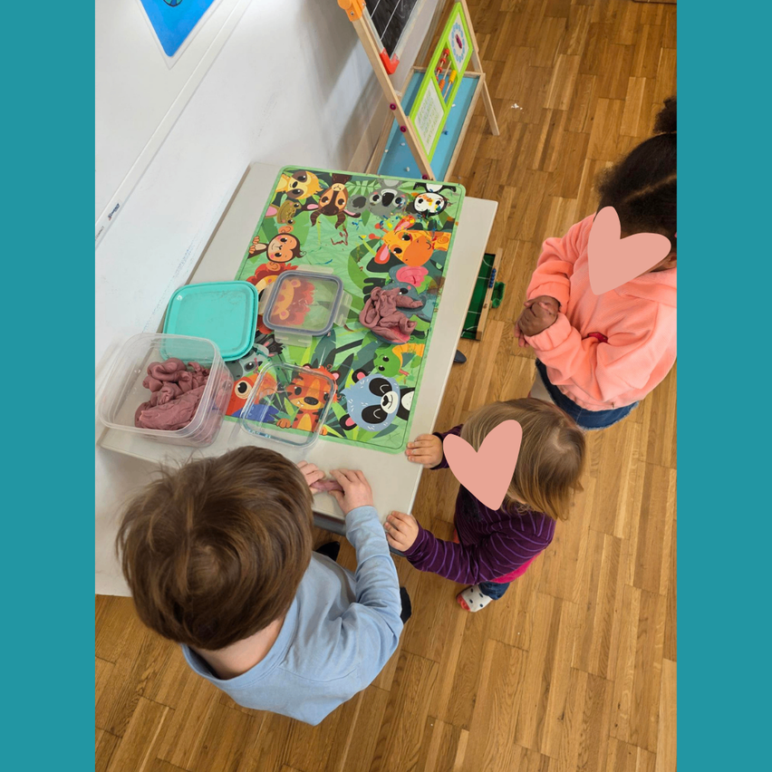 Drei Kinder spielen mit Ton auf einem Tisch mit einer tierthematischen Matte. Ein Kind sitzt und zwei stehen. Der Raum hat Holzböden und eine weiße Wand.