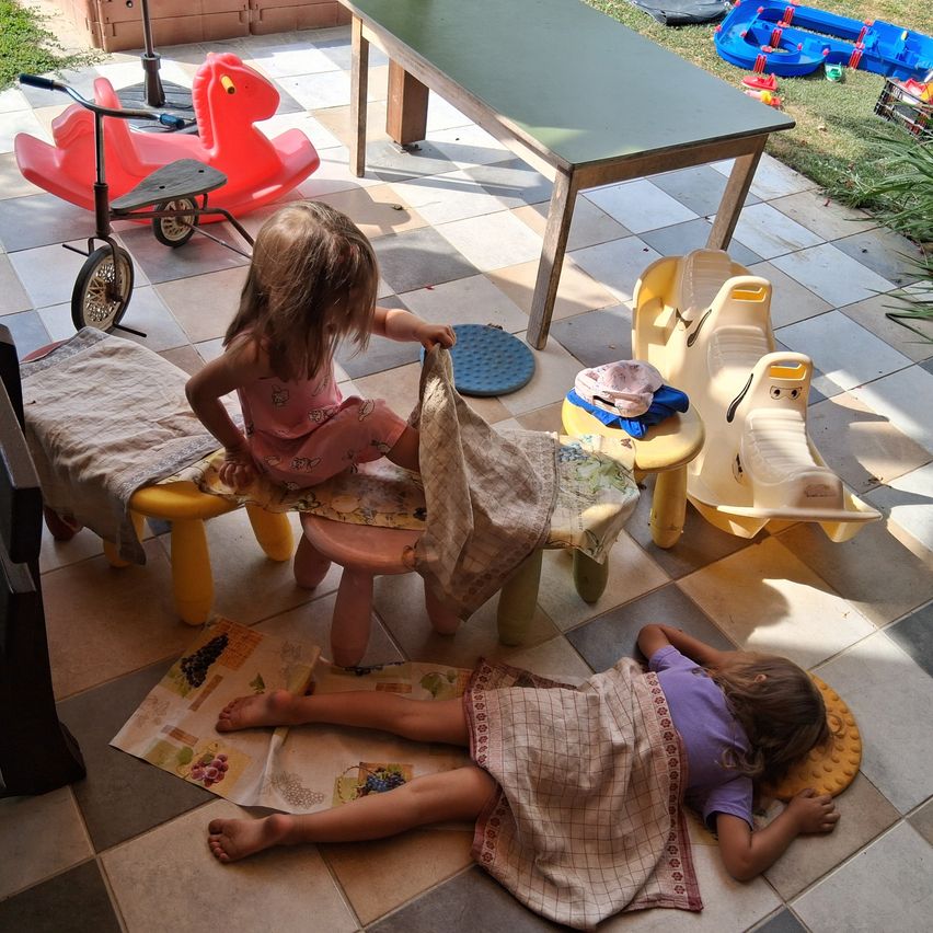 Zwei kleine Mädchen spielen auf der Terrasse mit Spielzeug. Eine sitzt auf einem Stuhl, hält ein Tuch, und die andere liegt auf dem Boden.
