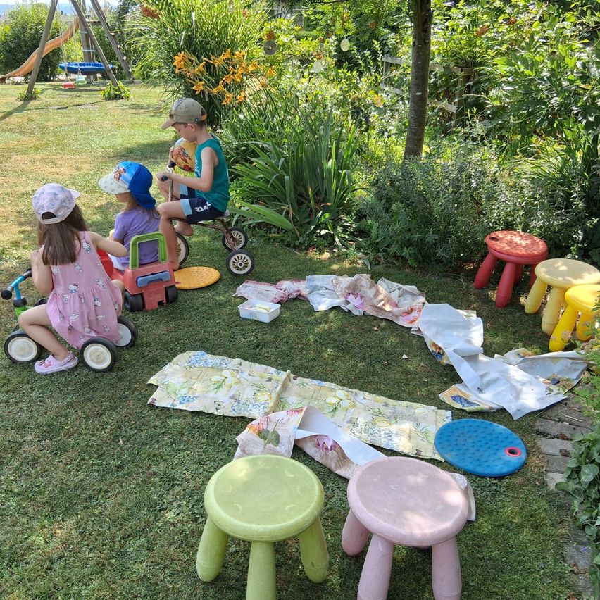 Drei Kinder spielen in einem Garten mit Spielzeug und bunten Stühlen. Ein Kind sitzt auf einem Dreirad, während die anderen auf einem Spielzeugauto sitzen.