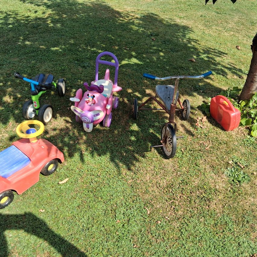 Vier Kinderspielzeuge stehen auf einem Rasen. Ein rosa Spielzeugflugzeug, ein grünes Dreirad, ein rotes Dreirad und ein rotes Spielzeugauto. Ein roter Benzintank steht rechts.