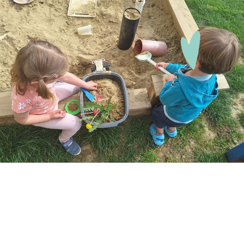 Zwei Kinder spielen in einem Sandkasten, eines gräbt mit einer Schaufel und das andere pflanzt Blumen.