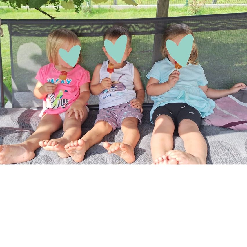 Drei kleine Kinder sitzen auf einer Bank, jedes hält eine Lutscher, mit herzförmigen Aufklebern auf den Gesichtern. Sie sind barfuß und in lässiger Sommerkleidung gekleidet.