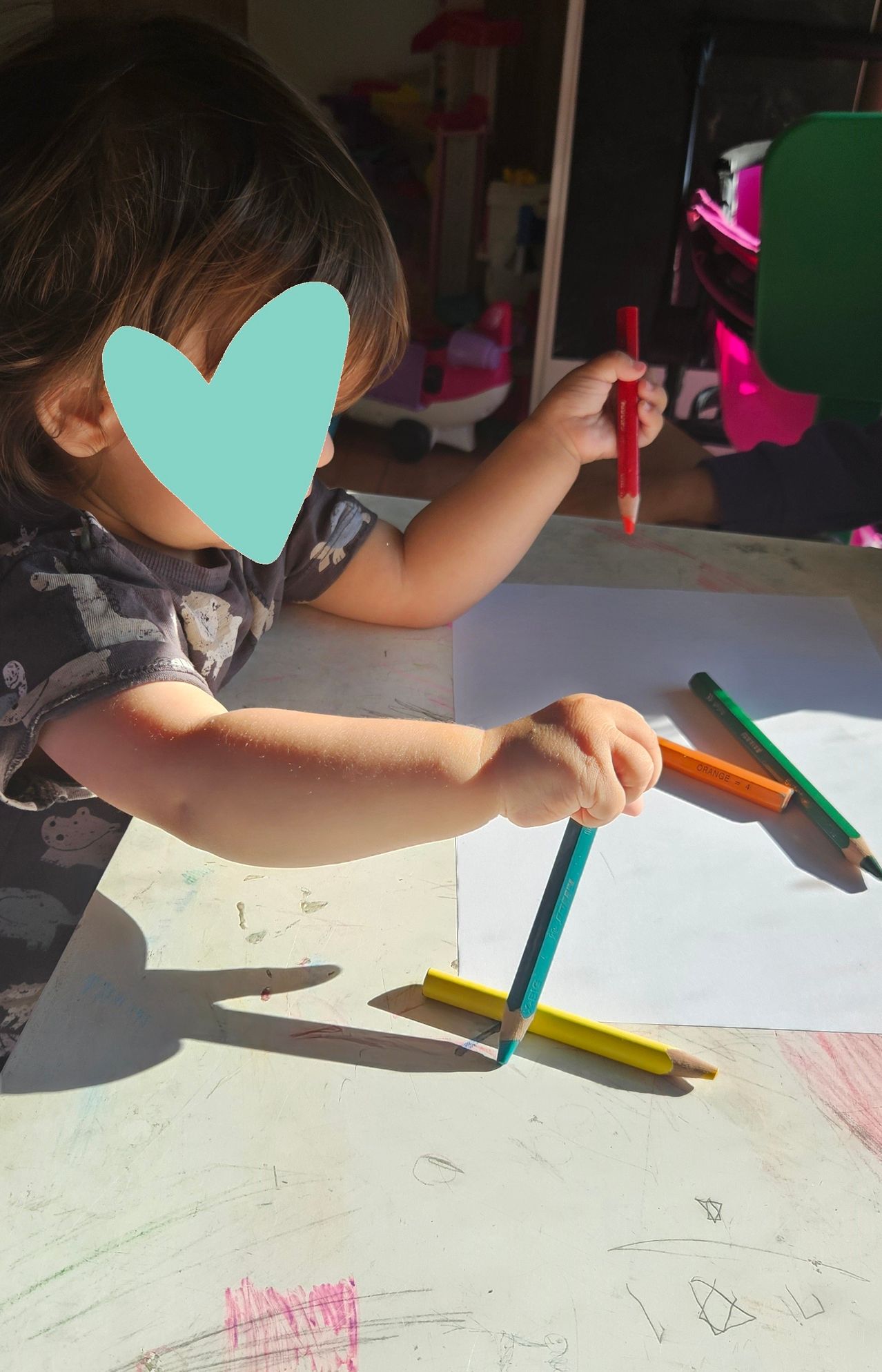 Ein Kind sitzt an einem Tisch und hält einen blauen Stift in der Hand, während es auf einem Blatt Papier zeichnet. Andere farbige Stifte liegen auf dem Tisch.
