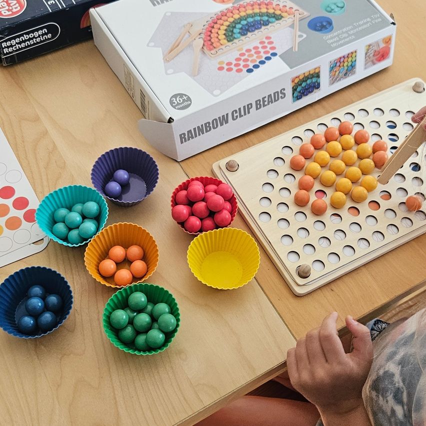 Ein Kind spielt mit einem Set von Regenbogen-Clip-Perlen und benutzt ein Brett mit Löchern und kleinen Bechern, die mit bunten Perlen gefüllt sind.