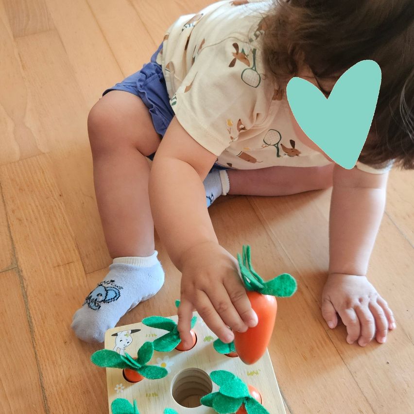 Ein Kleinkind spielt mit einem hölzernen Spielzeug, das Karotten zeigt, auf einem Holzboden sitzend. Das Kind trägt Socken mit einem Elefantendruck.