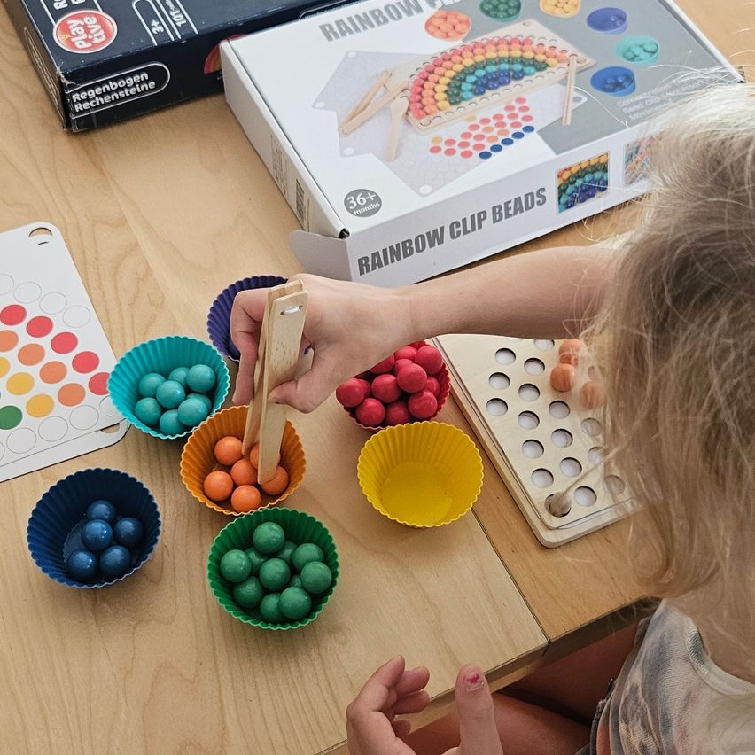 Ein junges Kind spielt mit einem Set von Regenbogen-Clip-Perlen und ordnet bunte Perlen in kleinen Bechern auf einem Holztisch an.