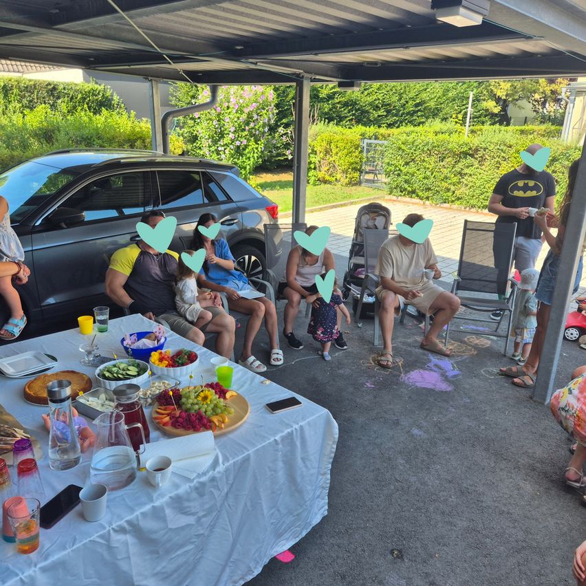A family gathering in a carport with a table full of food, drinks, and a baby in a stroller. Some adults are wearing face masks.