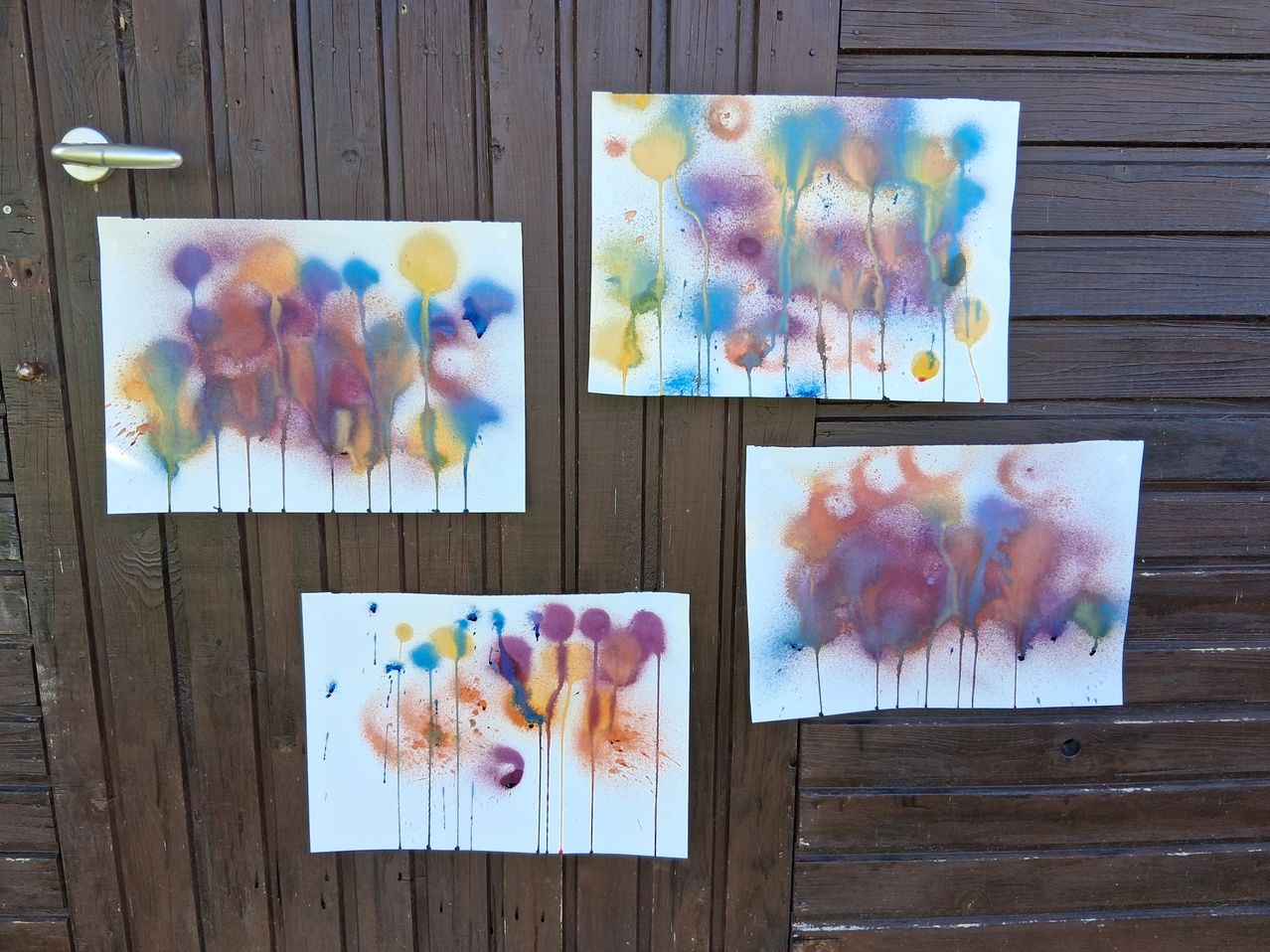Vier abstrakte Gemälde mit leuchtenden Farben und Spritzeffekten sind auf einer hölzernen Oberfläche ausgestellt. Jedes Gemälde zeigt eine einzigartige Kombination aus blauen, gelben, orangefarbenen und lila Farbtönen.