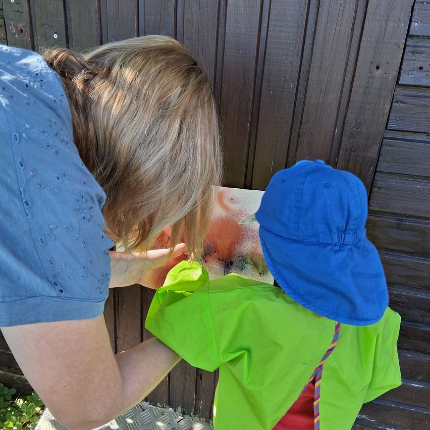 Eine Frau und ein Kind malen eine Holzwand mit leuchtenden Farben. Die Frau trägt ein blaues Shirt und das Kind einen grünen Regenmantel und einen blauen Hut.