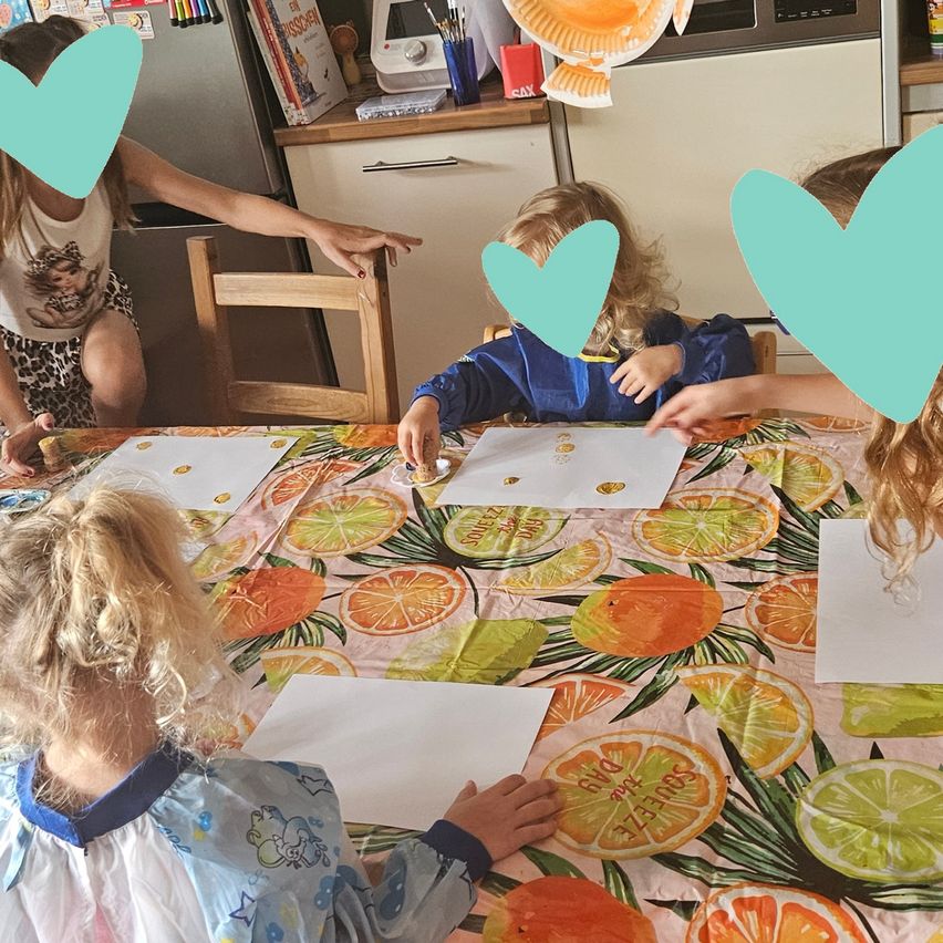 Four children are seated around a table covered with an orange and lemon-themed tablecloth. They are engaged in an activity, possibly painting or drawing, with papers and paintbrushes on the table. One child is wearing a blue smock.