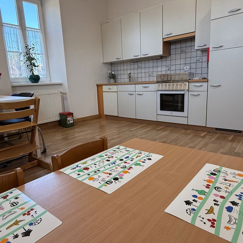 A clean, well-lit kitchen with wooden floors and a dining table set with colorful placemats. A window with a potted plant and a radiator is visible on the left.