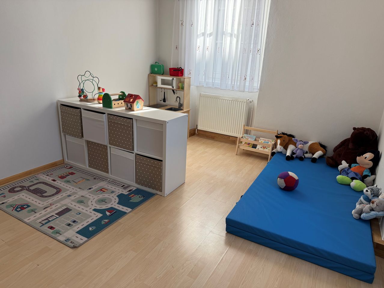 A child's playroom with a white shelf, toys, and a blue mat. A white radiator and a window with white curtains are in the background.
