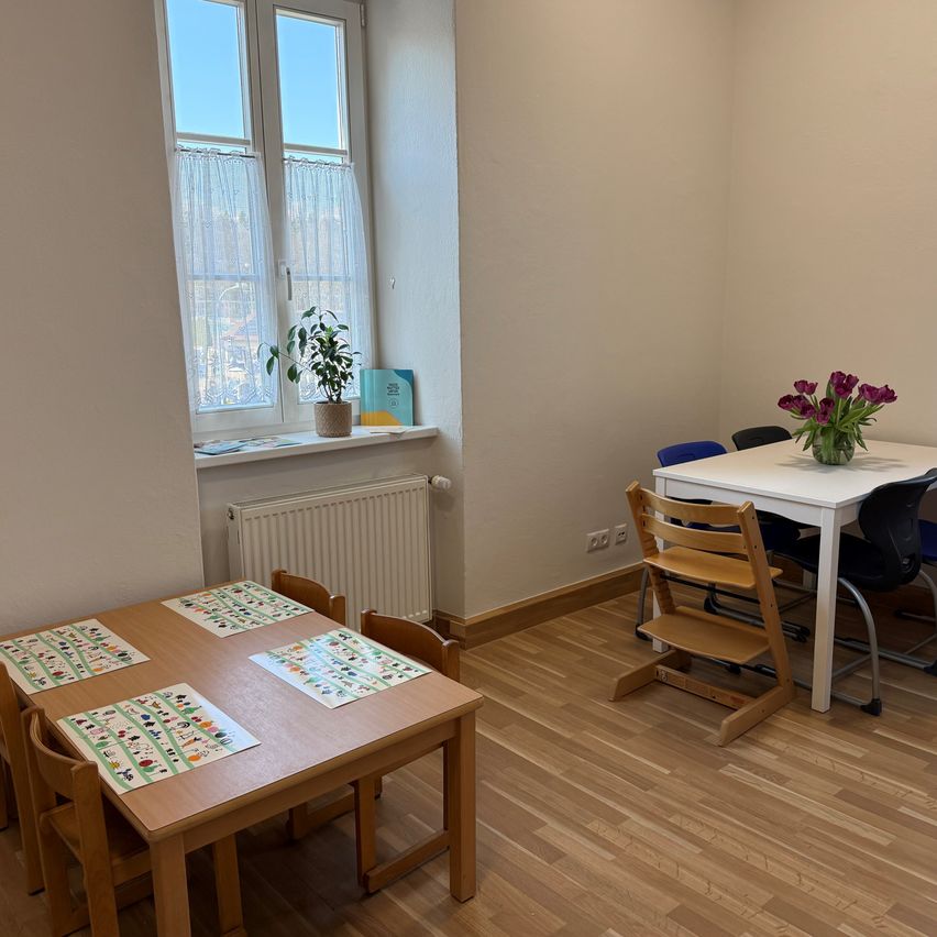 A room with wooden floors, a white wall, a window with a plant and book, and two tables with colorful placemats.