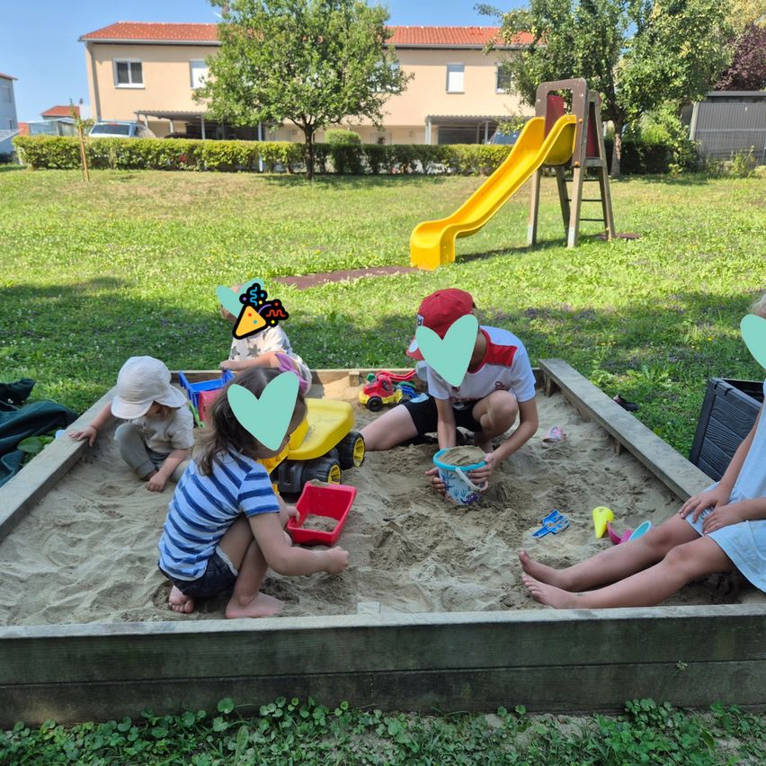 Four children play in a sandbox, one with a red bucket, another with a toy truck, and two others with hats. A yellow slide and a house are in the background.
