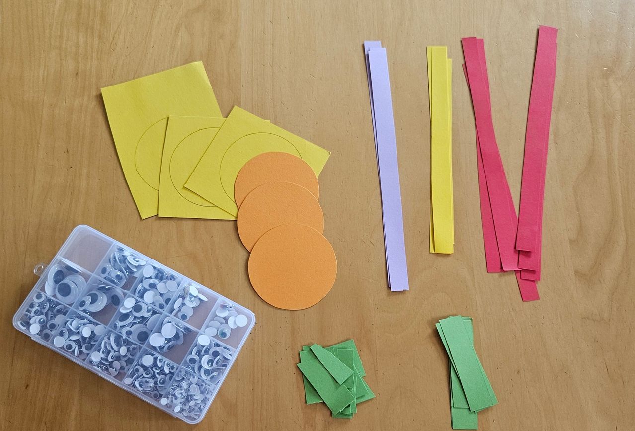 A wooden table displays various colored paper circles, strips, and a plastic container with assorted small items.
