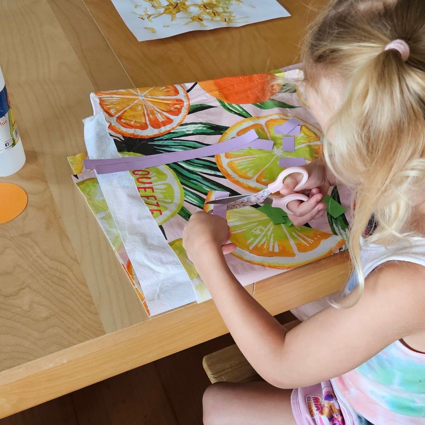 A young girl with blonde hair is cutting a piece of fabric with scissors. The fabric has a colorful design of fruits and leaves.