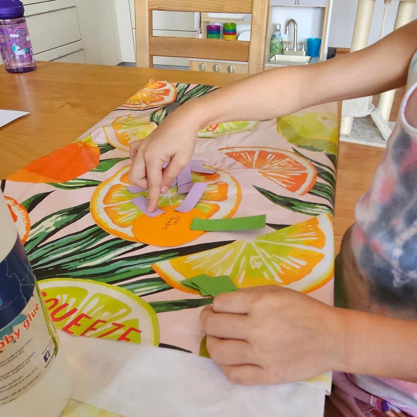 A child is sitting at a table, working on a craft project with paper cutouts of citrus fruits. A bottle of glue and a white cloth are nearby.