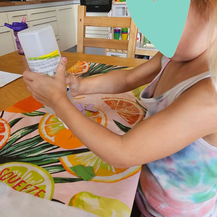 A young girl is sitting at a table, holding a bottle of glue and working on a craft project with orange and green paper cutouts.