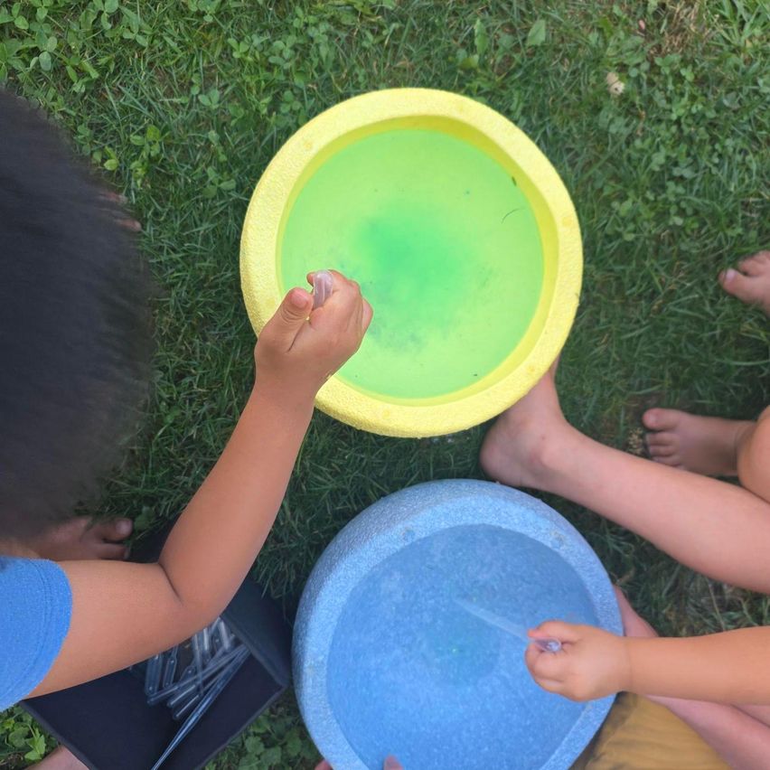 Two children are playing with water in yellow and blue basins on a grassy field. One child is adding green liquid to the yellow basin.
