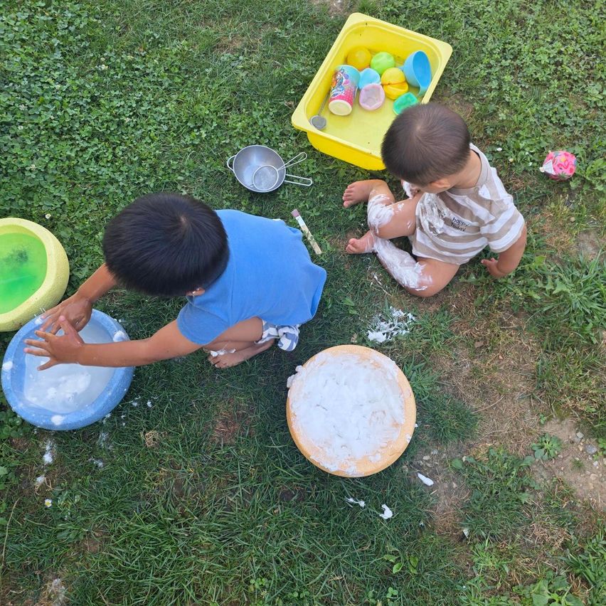 Two young boys are playing on the grass with water and foam. One boy is sitting and playing with foam while the other is standing and playing with water.