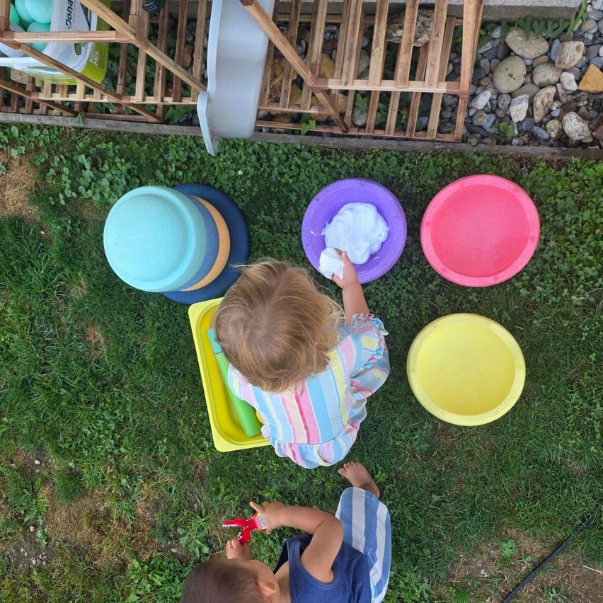 Two children are playing on the grass with colorful bowls and toys. One child is sitting and holding a bowl with foam, while the other child is standing and holding a toy.