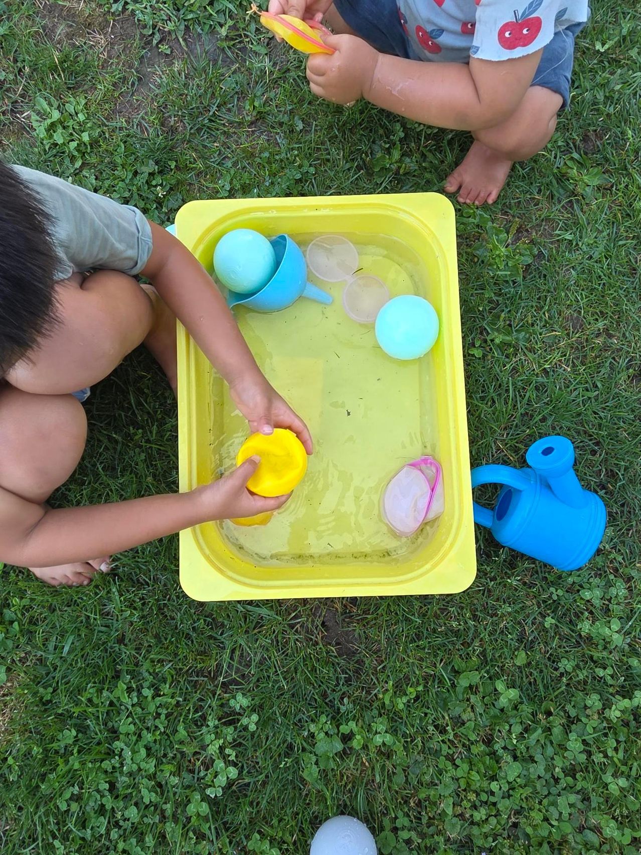 A child is playing with water and toys in a yellow tray on a grassy lawn. The child is holding a yellow ball and there are other toys in the tray.