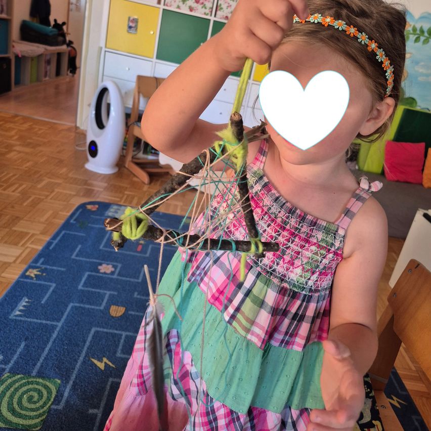 Ein kleines Mädchen in einem bunten Kleid hält einen Stock mit einem herzförmigen Aufkleber. Sie steht in einem Zimmer mit einem blauen Teppich und einem weißen Ventilator.