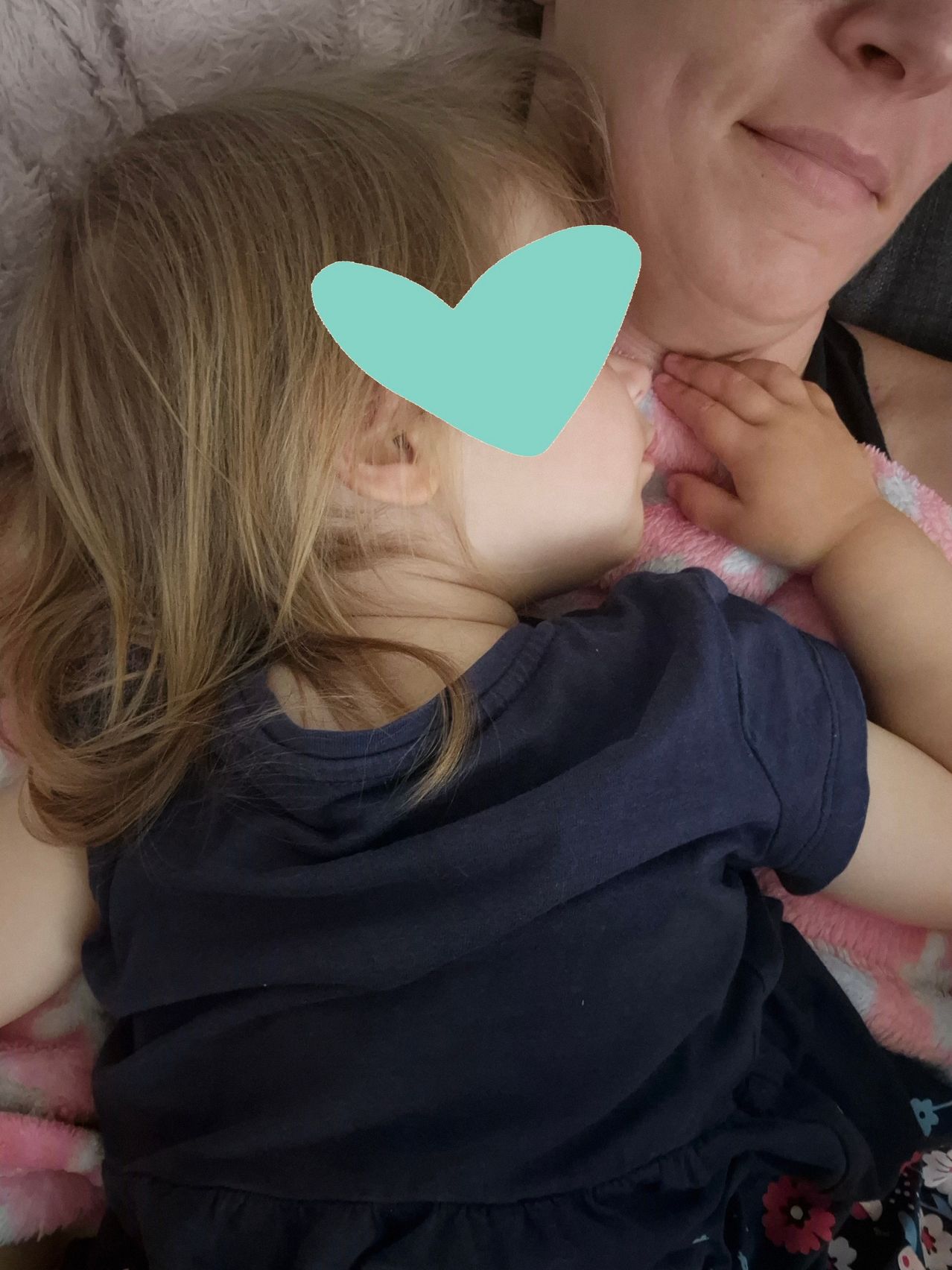 Ein kleines Mädchen in einem dunklen blauen T-Shirt liegt auf einer rosa Decke und küsst eine Frau, die sie hält. Die Hand der Frau liegt auf der Wange des Mädchens.