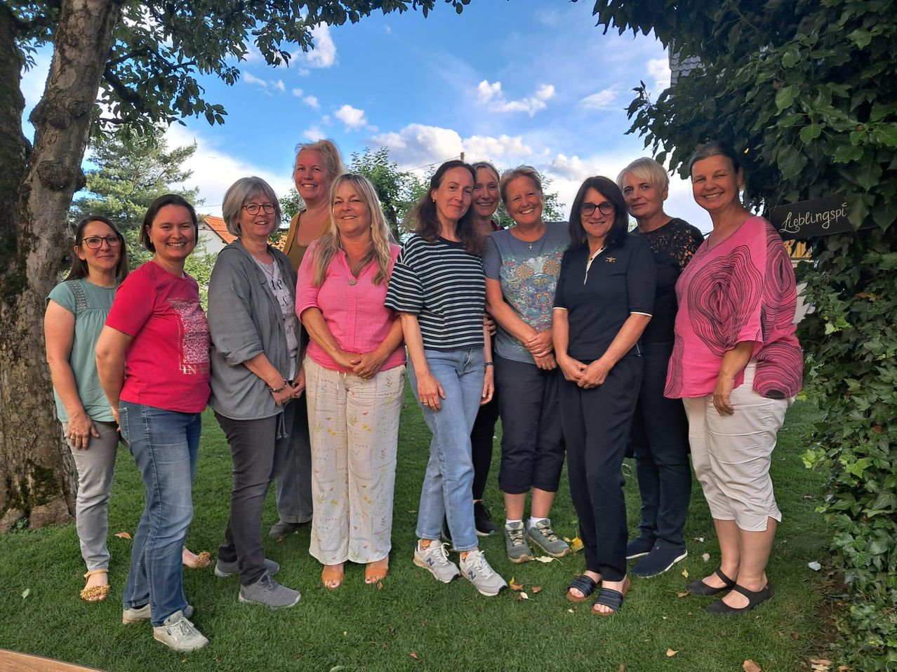 Eine Gruppe von Frauen in Freizeitkleidung, die lächelnd in einem Garten stehen. Sie sind in einem Halbkreis angeordnet, mit Bäumen im Hintergrund.
