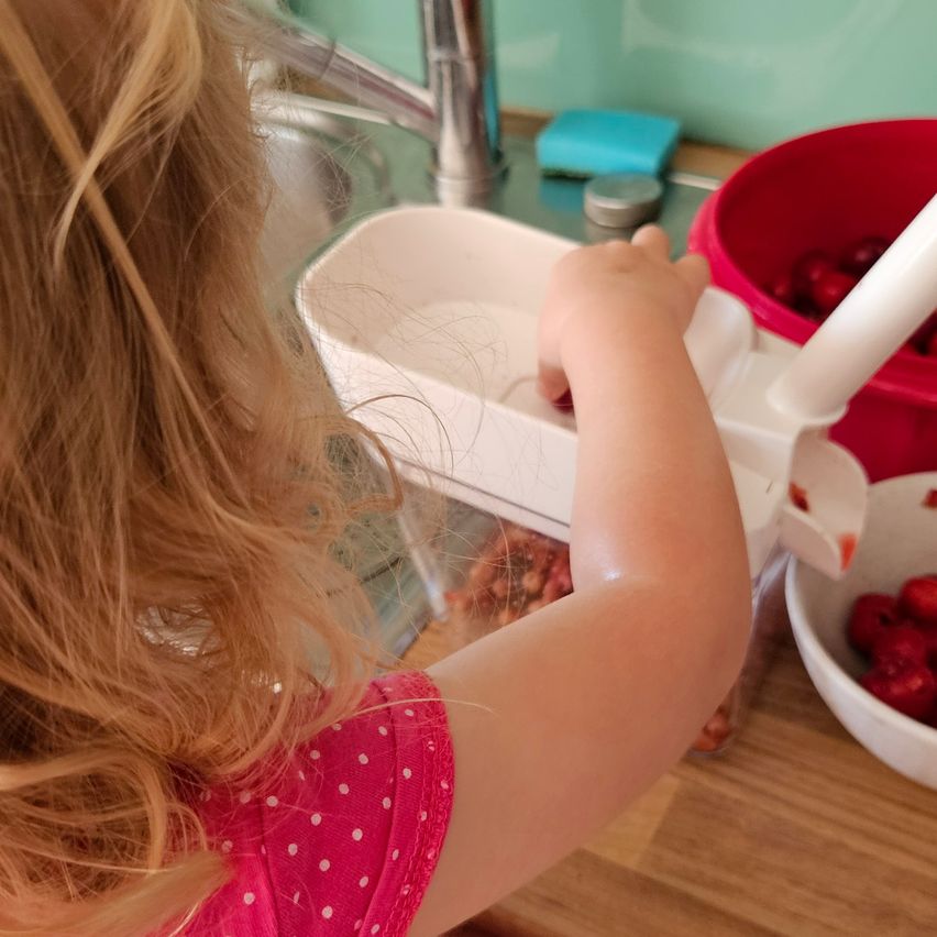 Ein junges Mädchen benutzt in der Küche einen Erdbeerschneider, mit Erdbeeren in einem roten Eimer und einer weißen Schüssel in der Nähe.