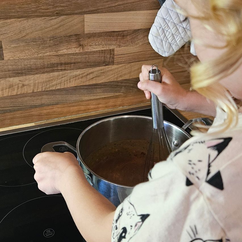 Ein junges Kind rührt in einem Topf Sauce auf dem Herd, während es eine Schlagsahne hält. Die Küche hat Holzwände.